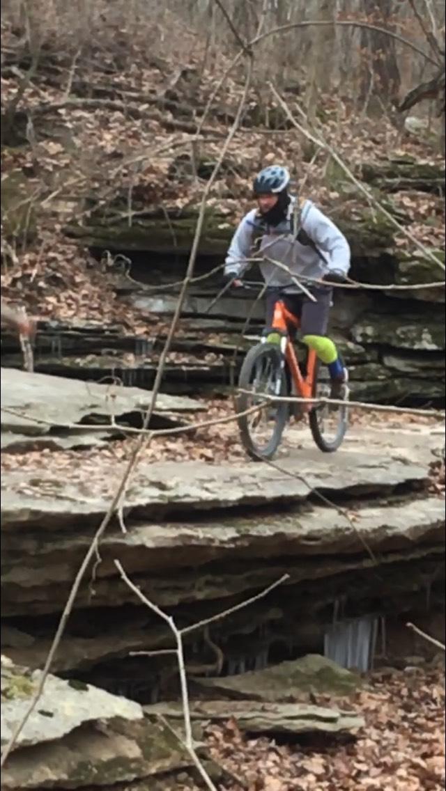 A mountain biker is navigating over rocky terrain in a forested area, surrounded by fallen leaves and tree branches. The biker is wearing a helmet and is dressed in a light gray jacket and shorts, with bright green knee pads. Various layers of rock are visible beneath the biker's wheels as they ride on a trail. Back 40 mountain bike trail.