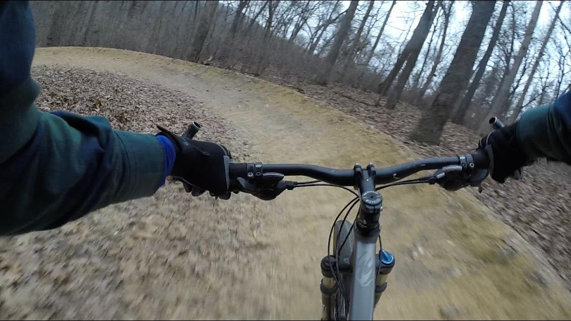 A close-up view of a mountain bike's handlebars as it navigates a winding dirt trail lined with fallen leaves in a wooded area. The rider's gloved hands grip the handlebars, indicating an active cycling experience. The scene captures a sense of motion and adventure in a natural setting during a cool, overcast day. Coler Mountain Bike Preserve mountain bike trail.