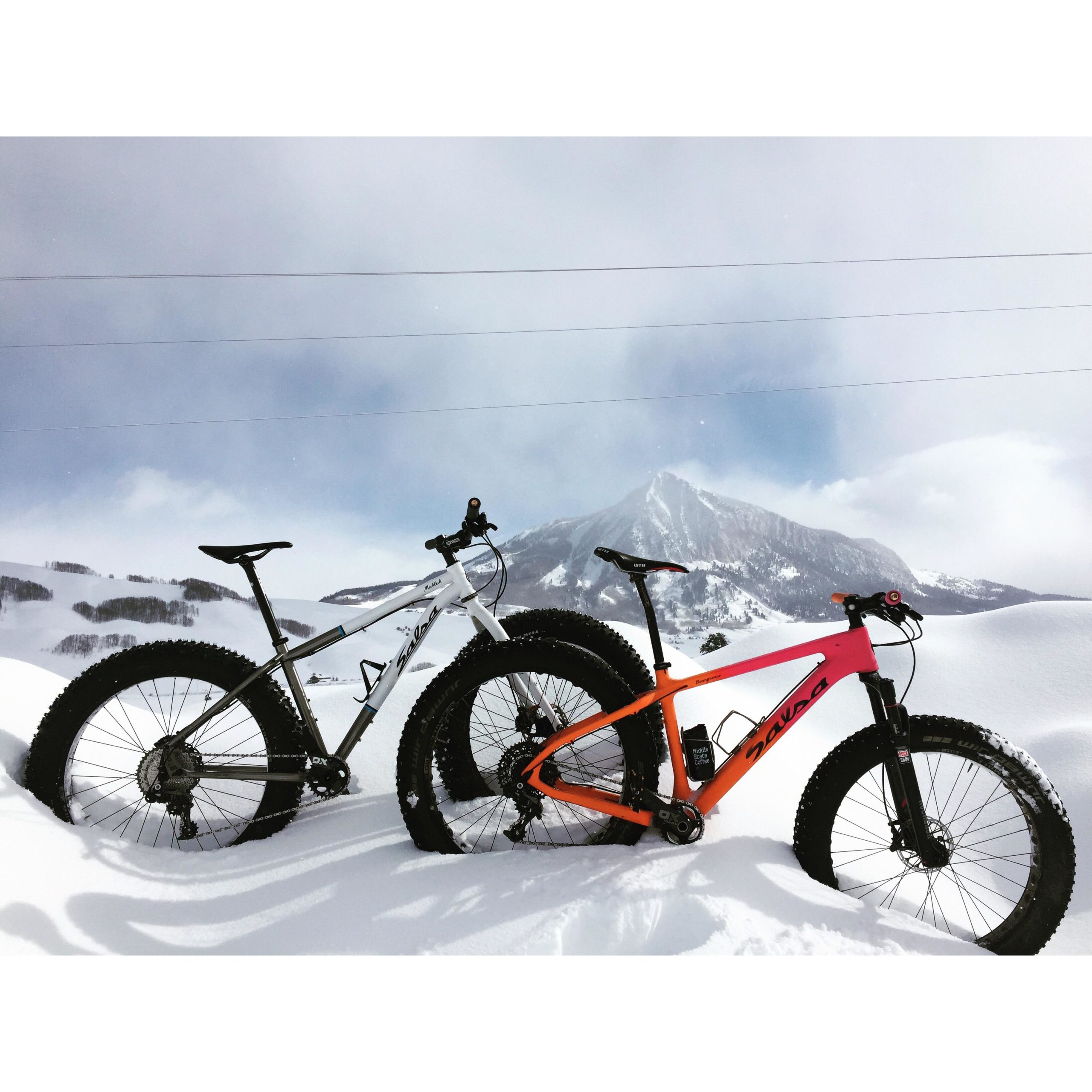 Two fat bikes, one white and one orange, are parked in deep snow with a mountain backdrop. The scene captures a winter landscape with a cloudy sky and snow-covered hills. Arcadia Laketrails mountain bike trail.