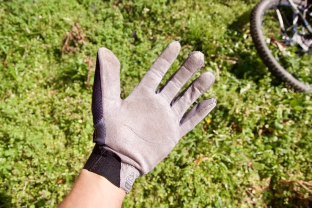 A close-up view of a gray glove with a textured palm, held up against a backdrop of green grass and vegetation. A bicycle wheel is slightly visible in the background.