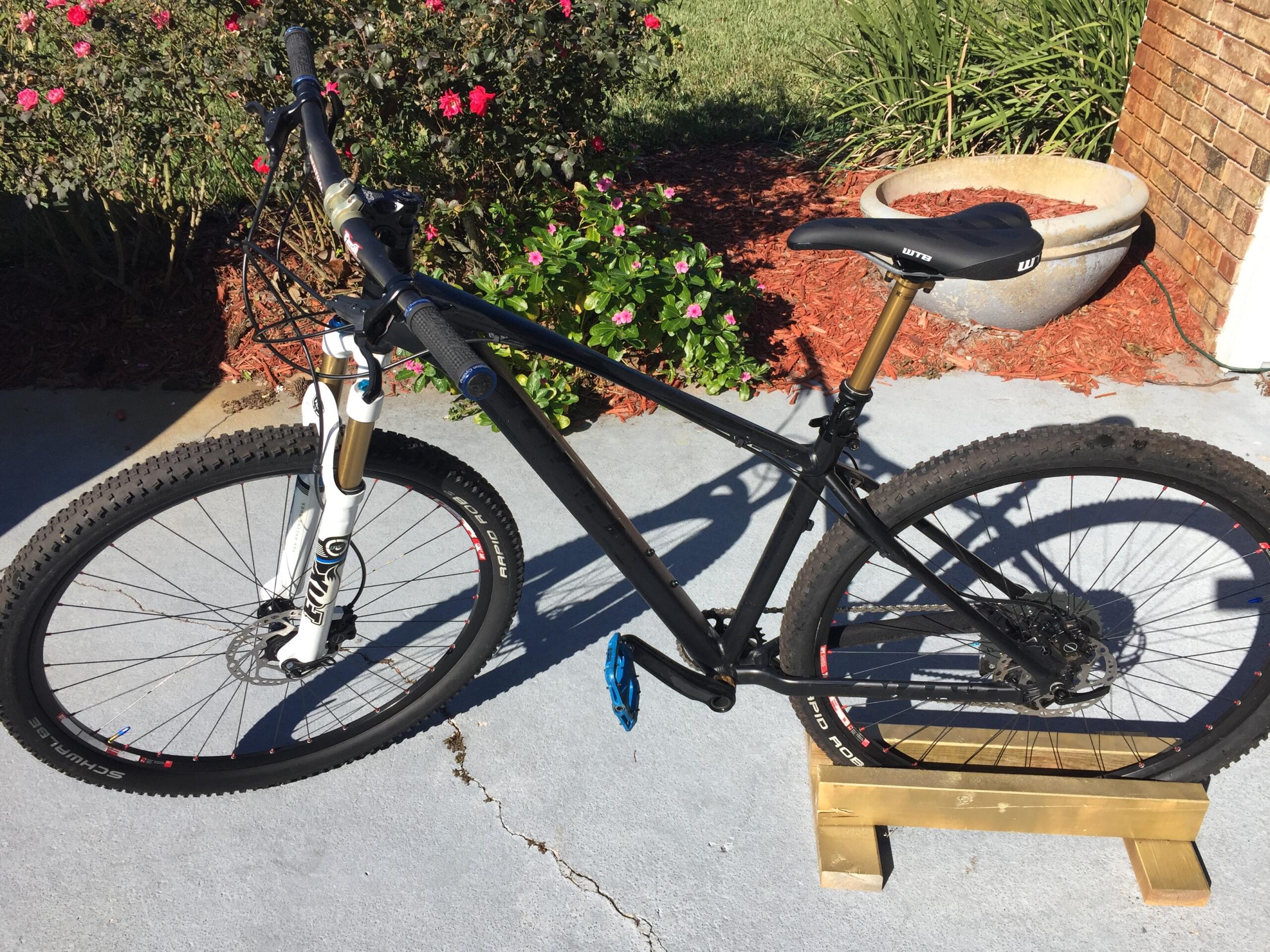 Giant Talon 29er: A black mountain bike with a white front fork is positioned on a wooden stand. The bike features thick, knobby tires and a sleek design, with a prominent seat and handlebars. In the background, vibrant pink flowers and green shrubs create a colorful outdoor setting. Sunlight casts a clear shadow on the concrete surface.