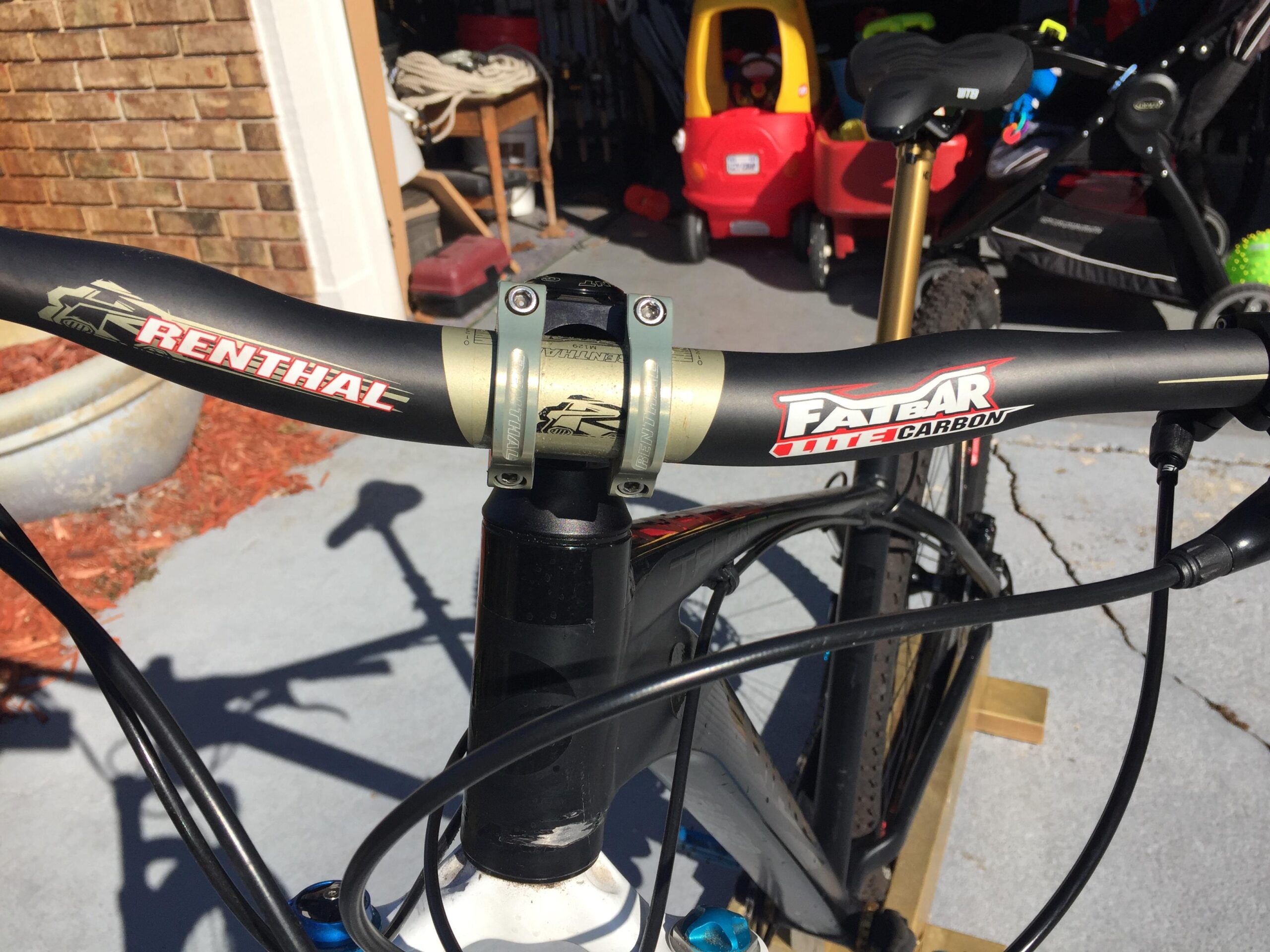 Giant Talon 29er: Close-up view of a mountain bike's handlebars featuring "Renthal" and "Fatbar Lite Carbon" branding, with a visible headset and cables. The background includes a garage setting with various outdoor items and a toy vehicle.