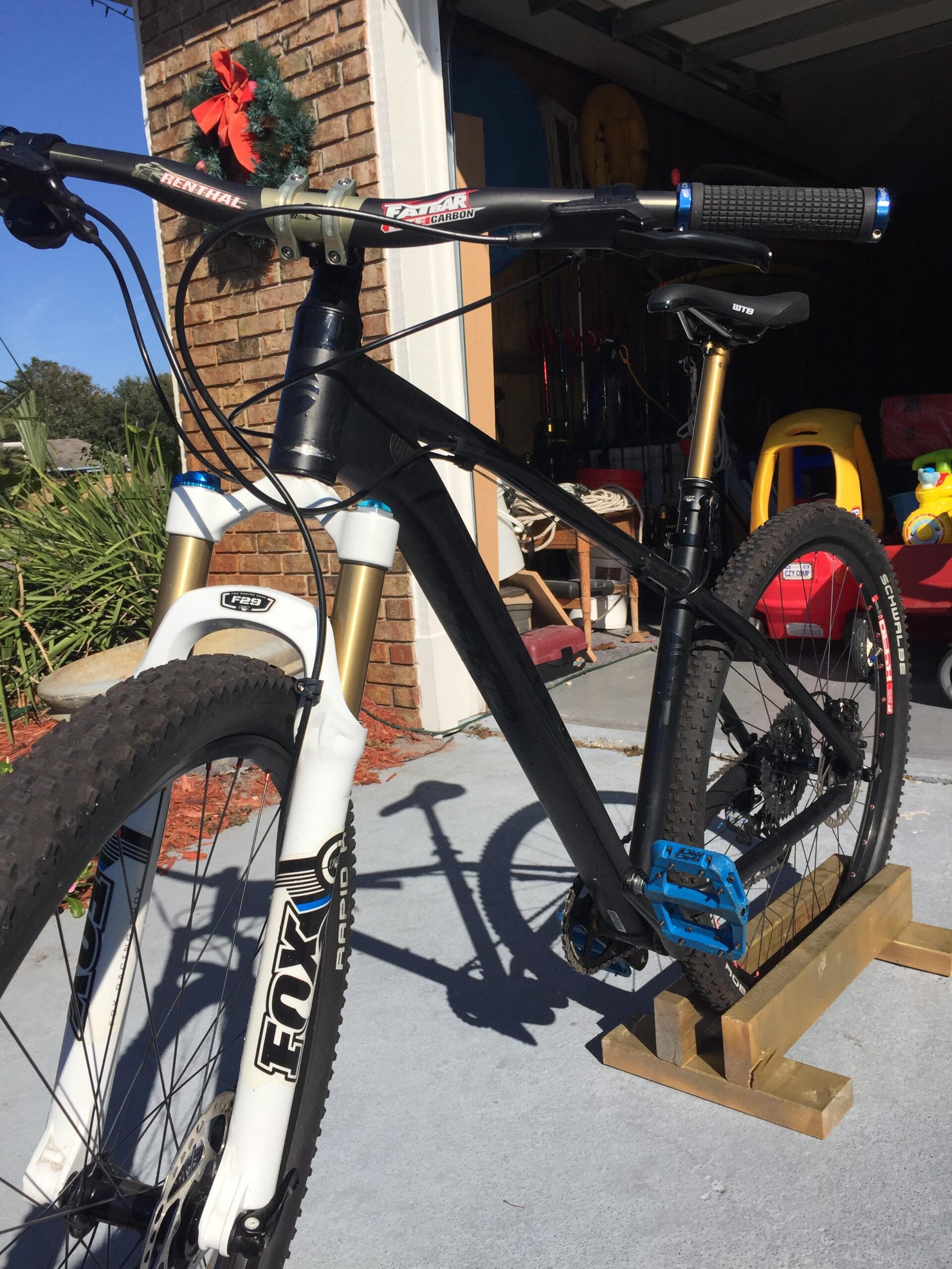Giant Talon 29er: A black mountain bike with a white and gold front fork, resting on a wooden bike stand. The handlebars feature black grips and a blue accent, and the bike has blue pedals. The background shows a garage with various items visible, including a wreath on the wall and children's toys.