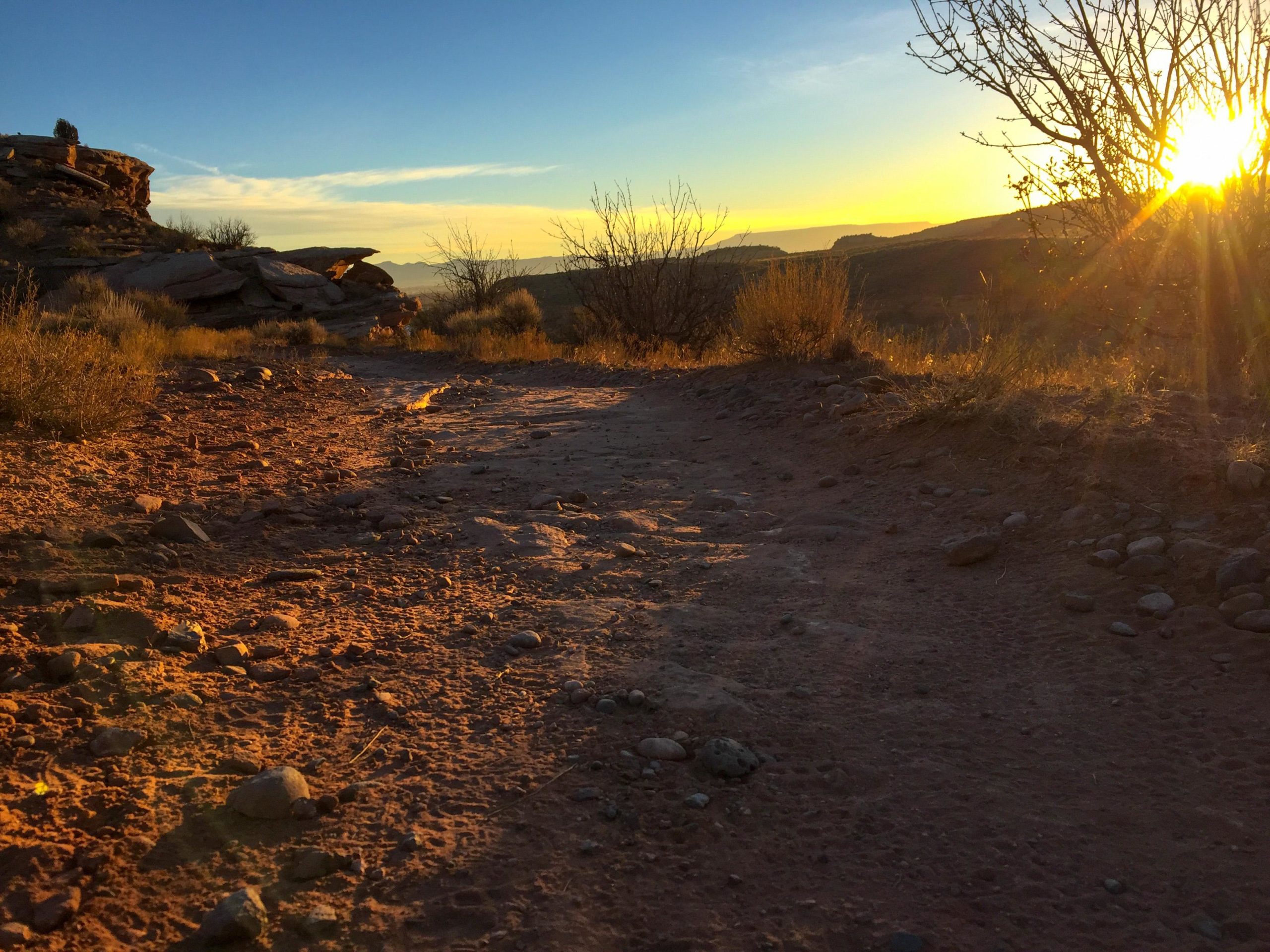 A dirt path leading through a rocky landscape at sunset, with the sun illuminating the scene and casting long shadows. Sparse vegetation and bushes are visible along the path, and distant hills can be seen in the background under a colorful sky. Kokopelli Trail mountain bike trail.