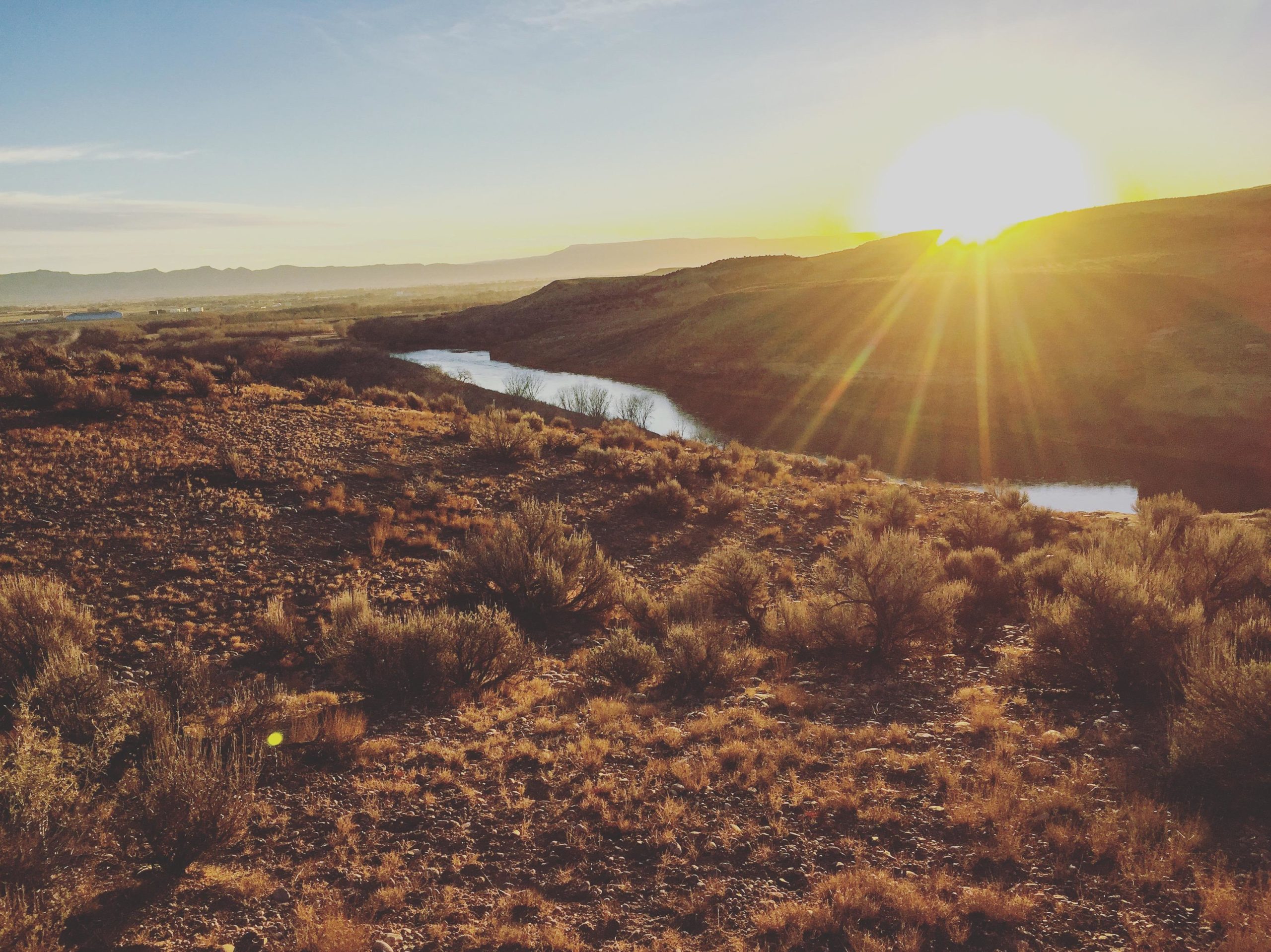 A stunning sunrise over a river surrounded by hills and sparse vegetation. The sunlight casts a golden hue across the landscape, highlighting the contours of the terrain and creating a warm, tranquil atmosphere. Kokopelli Trail mountain bike trail.