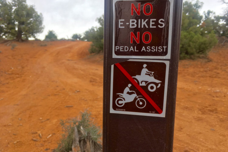 A sign post in a natural landscape, featuring two signs: one stating "NO E-BIKES NO PEDAL ASSIST" and another with symbols indicating a prohibition on motorcycles and ATVs. The background shows a dirt path and vegetation typical of a desert environment.