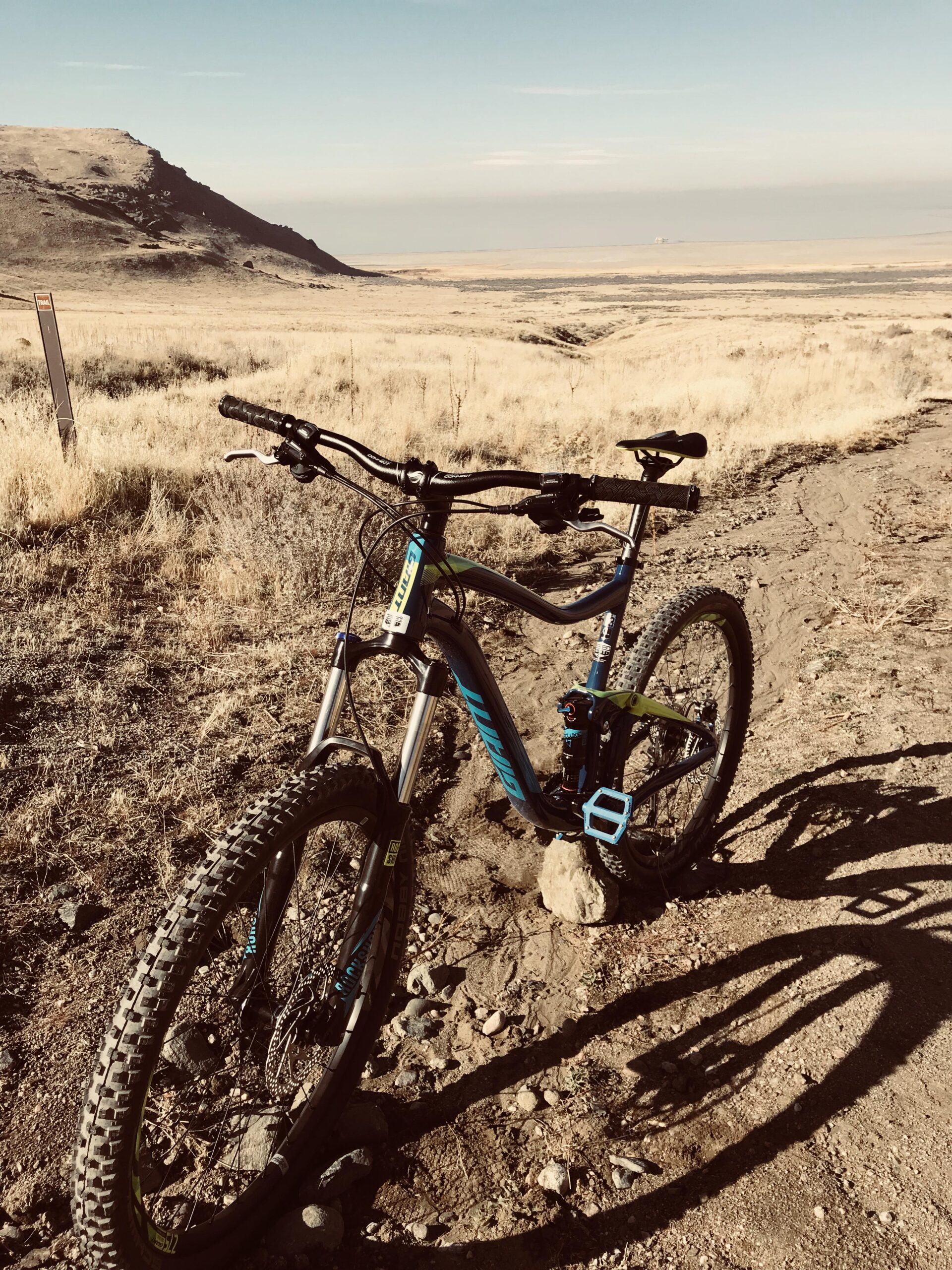 Giant Trance: A mountain bike resting on a dirt trail with grass and rocky terrain, surrounded by open fields and distant mountains under a clear blue sky. The bike features black and blue colors with visible shadows cast on the ground.