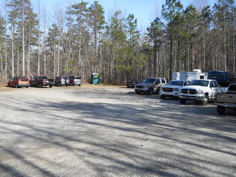 A gravel parking lot with several parked vehicles, including trucks and SUVs, set against a backdrop of trees. A portable toilet is visible in the background. Jake Mountain Trails mountain bike trail.