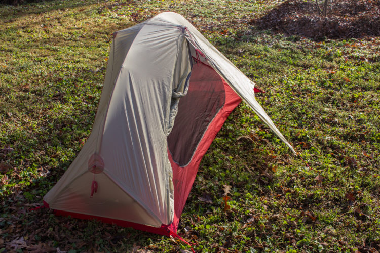 A tent set up on a grassy area, featuring a light beige exterior and red accents. The tent has a sloped roof and an open side, revealing a mesh panel for ventilation. Surrounding the tent is a blend of green grass and fallen leaves, indicating an outdoor camping setting.