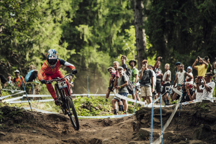 A mountain biker in a red and blue jersey navigates a dirt trail during a downhill race, surrounded by lush greenery. Spectators in the background cheer and take photos as the rider maneuvers around obstacles marked by racing tape. The scene captures the excitement and energy of a biking competition.