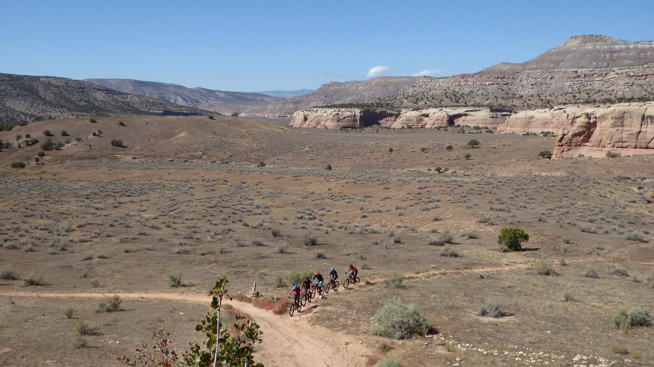 A group of mountain bikers riding along a dirt trail in a scenic desert landscape, with rolling hills and rocky cliffs in the background under a clear blue sky. Sparse vegetation and shrubs dot the terrain. Kokopelli Area Trails mountain bike trail.