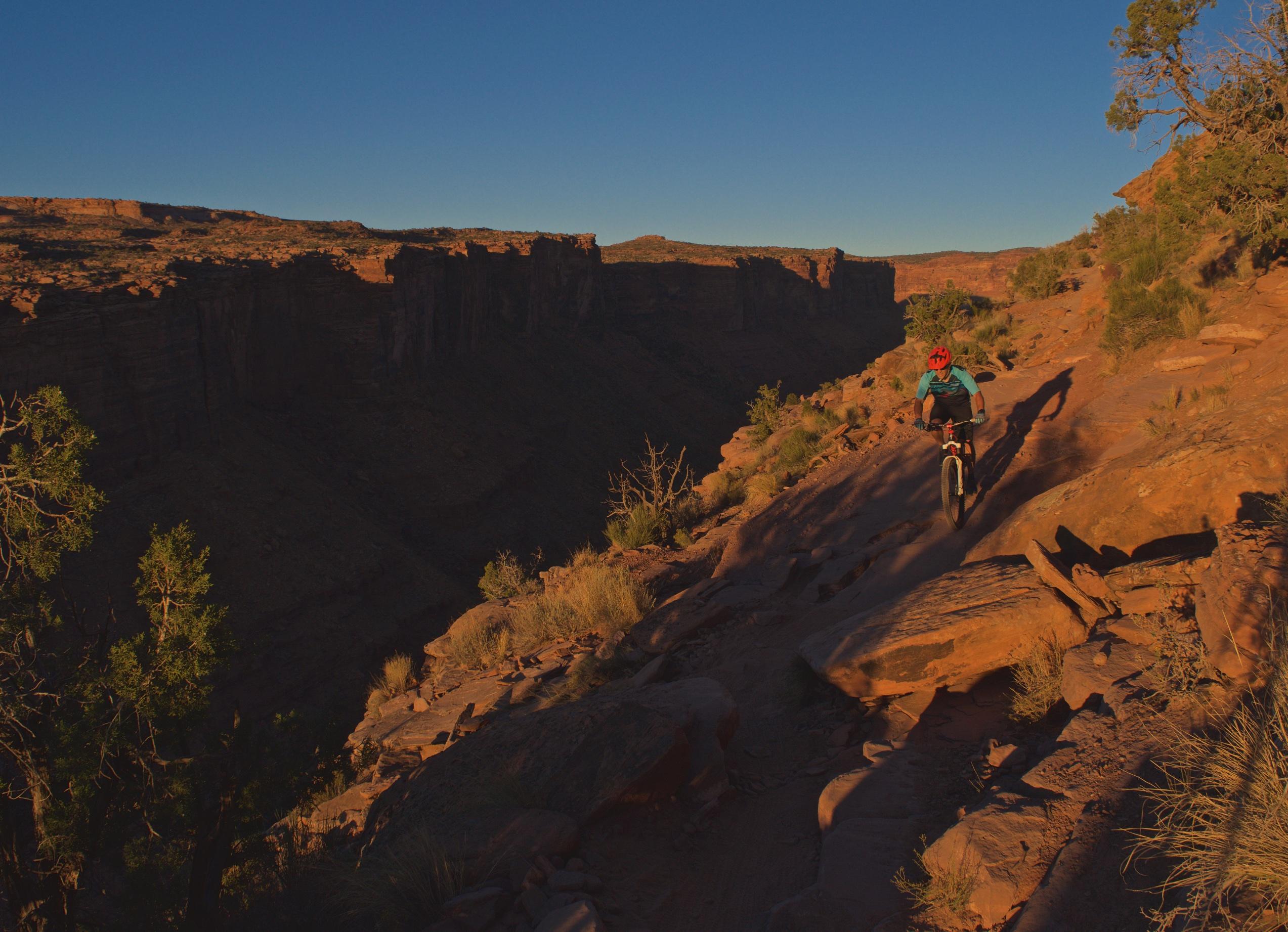 A mountain biker rides along a rocky trail in a canyon during golden hour, with dramatic cliffs in the background and a clear blue sky. The warm light casts long shadows on the terrain, highlighting the rugged landscape. Porcupine Rim mountain bike trail.