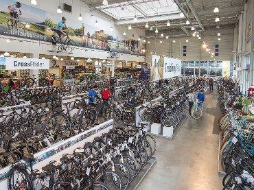 A spacious interior of a bicycle store featuring a wide selection of bicycles arranged on display. The store is well-lit with high ceilings and banners hanging above showcasing various bike categories. Customers are browsing the aisles, and there are multiple rows of bikes, including road bikes and mountain bikes.