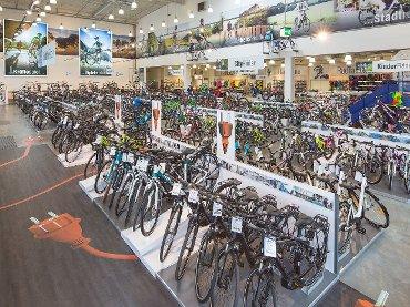 An expansive indoor bicycle store featuring a wide variety of bikes on display. Rows of bicycles are arranged neatly on the floor, with some prominently labeled, amidst bright lighting and vibrant promotional banners in the background.