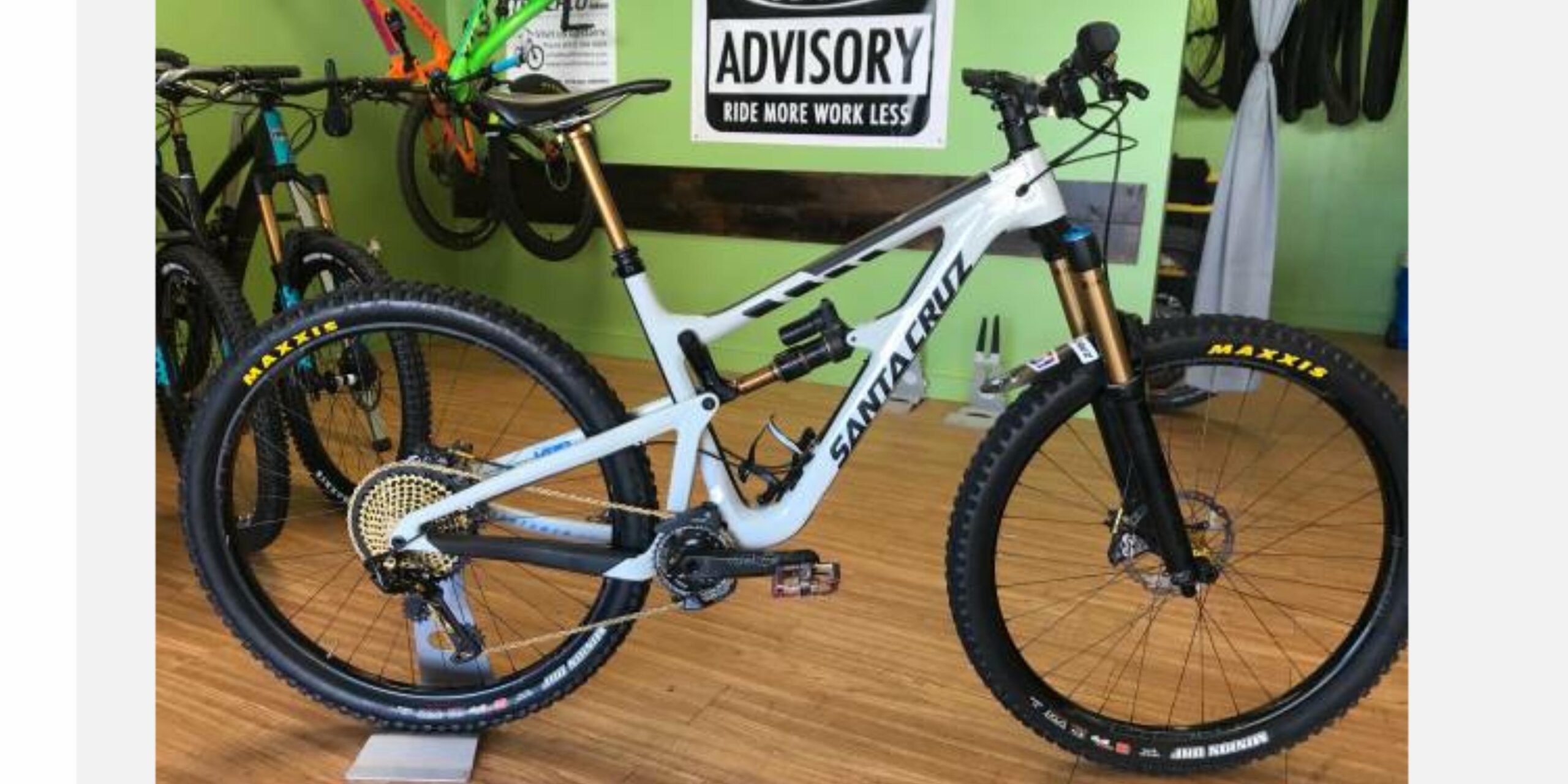 Santa Cruz Hightower 29er/27.5: A mountain bike displayed in a bike shop, featuring a white frame with black and gold accents. The bike has thick Maxxis tires and a modern design, showcasing its advanced suspension system. In the background, other bikes are visible along with a green wall that has an advisory sign reading "RIDE MORE WORK LESS."