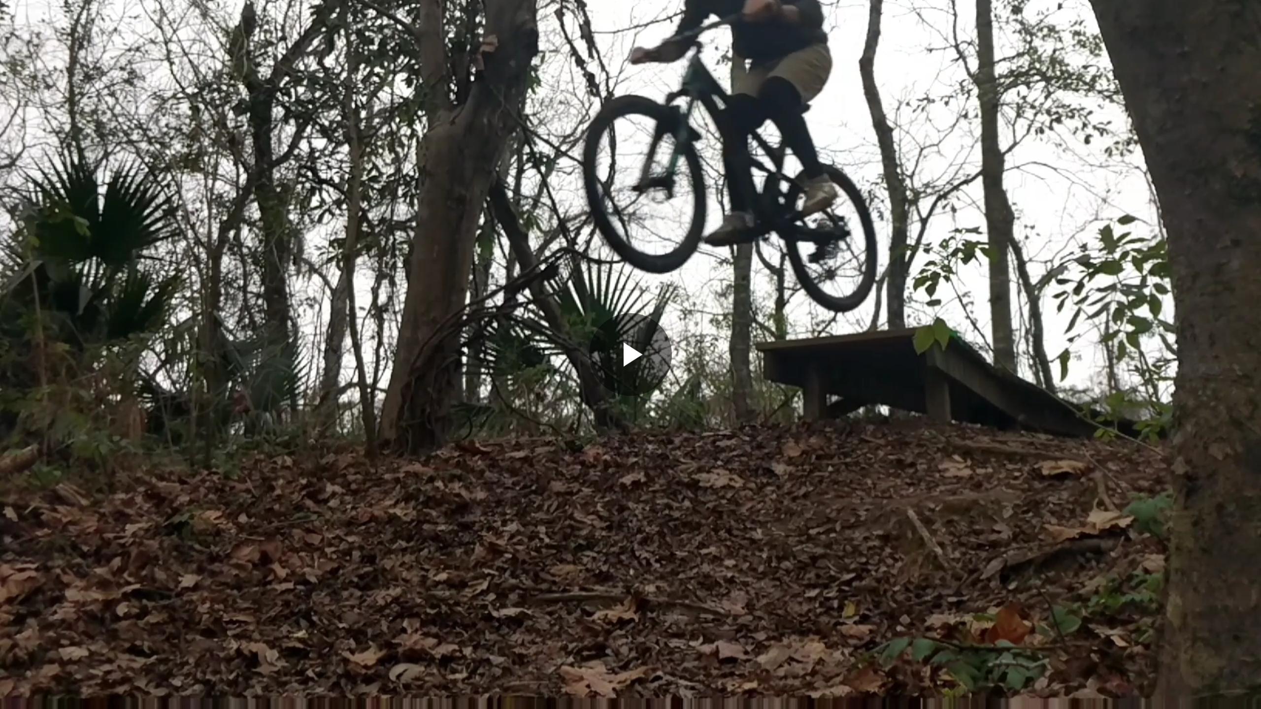 A person riding a mountain bike performs a jump off a small ramp surrounded by trees and fallen leaves. The biker is airborne, with the bike elevated above the ground, showcasing an adventurous moment in a natural outdoor setting. Bonnet Carre Spillway Trail mountain bike trail.