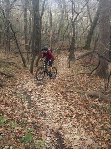 A person riding a mountain bike along a narrow dirt trail covered in autumn leaves, surrounded by trees in a wooded area. Lakewood Forest (Snowshoe / Snowmobile) mountain bike trail.