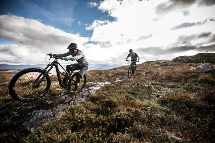 Two mountain bikers navigate a rocky trail in a scenic landscape. One rider is airborne, balancing on the front wheel while the other follows on a path lined with grass and stones beneath a dramatic sky.