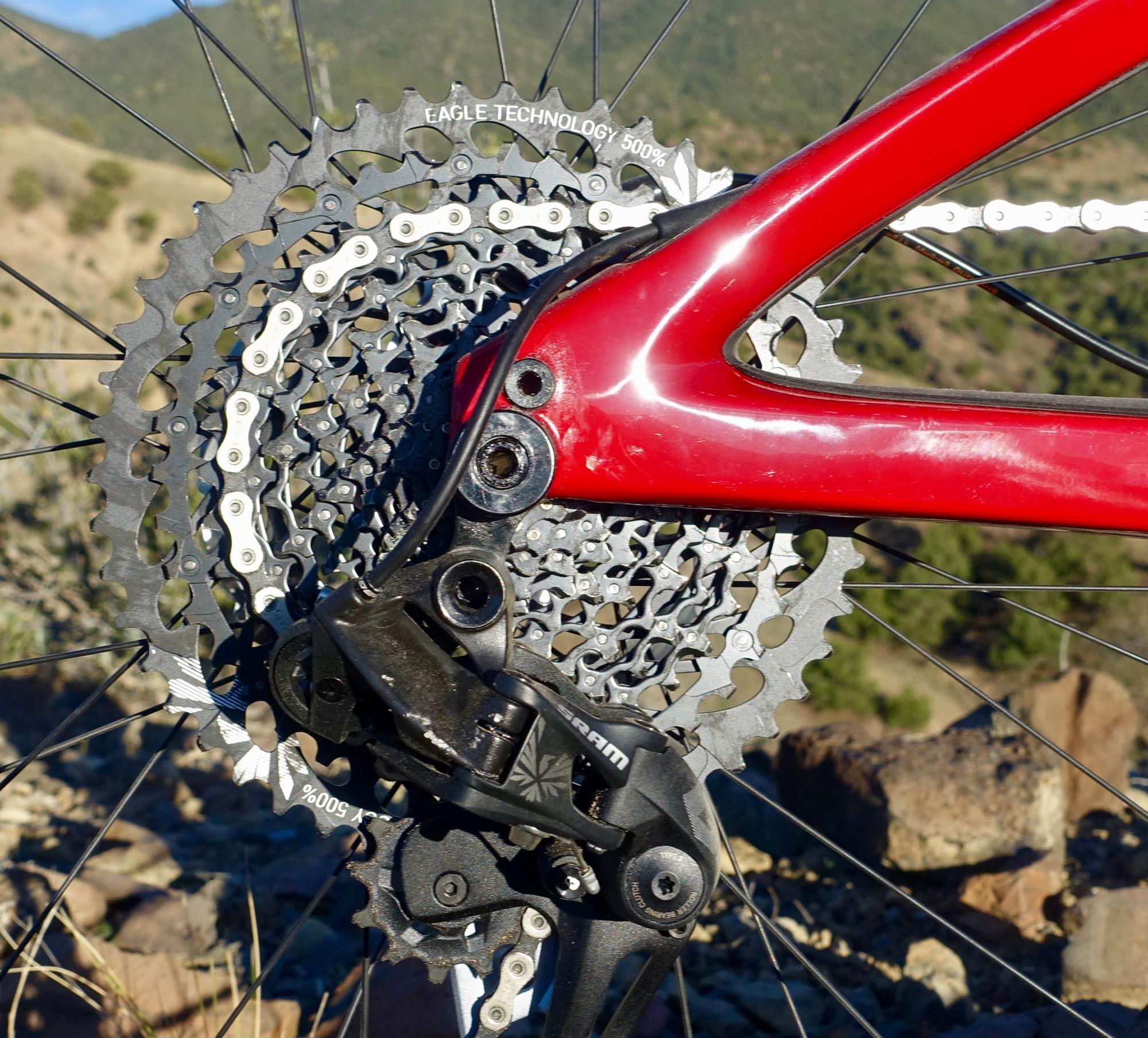 Ibis Mojo HD4: Close-up of a red bicycle's rear cassette and derailleur, showcasing the intricate gears and chain links with a focus on eagle technology branding. The background features a natural landscape with hills and rocky terrain.