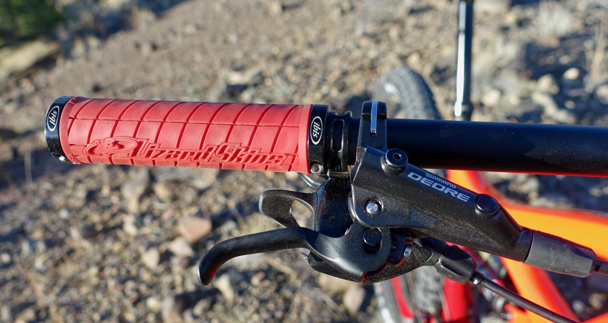 Ibis Mojo HD4: Close-up of a mountain bike handlebar showing a red rubber grip with textured surface and the brand name "Lizard Skins" embossed on it. The handlebar also features a Shimano Deore brake lever, with a blurred background of rocky terrain and a bike frame peeking through in orange.