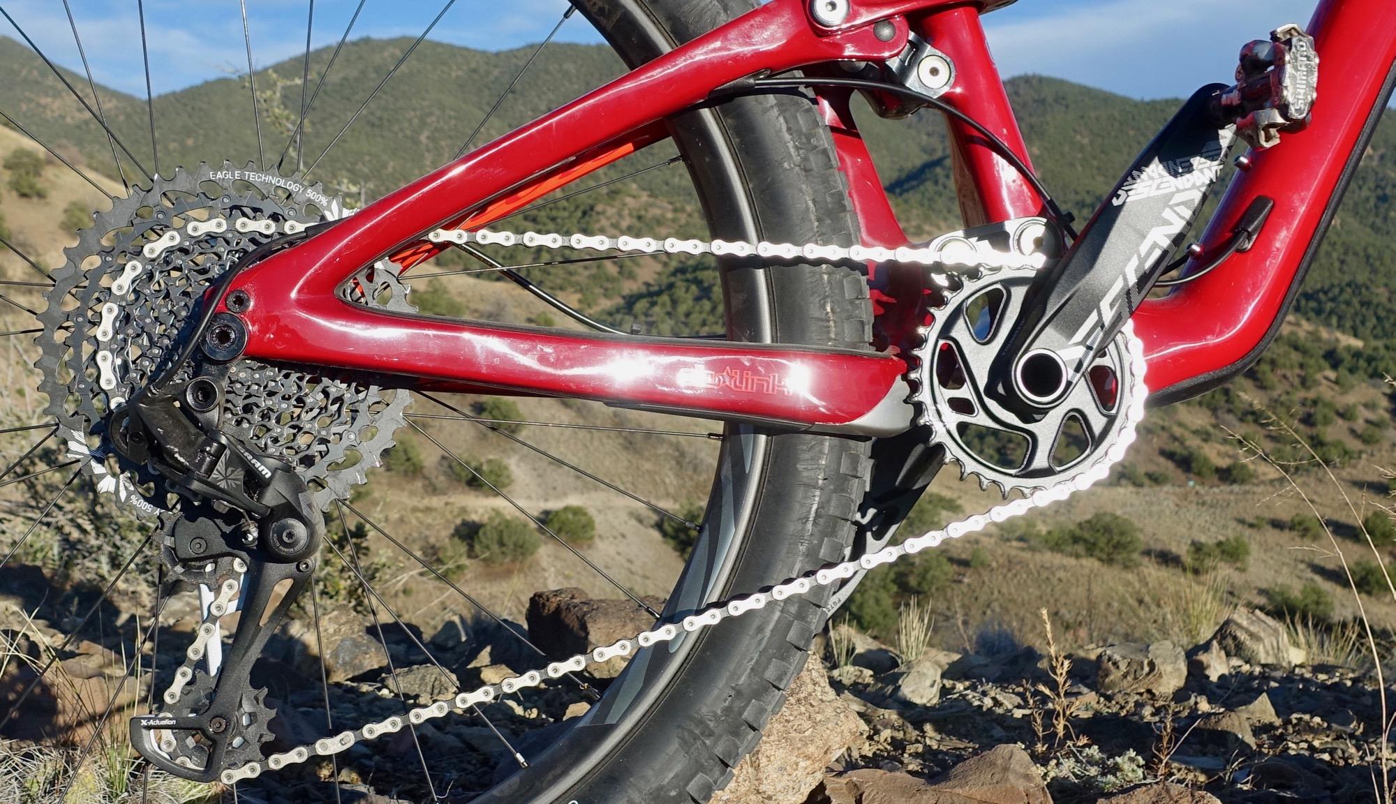 Ibis Mojo HD4: A close-up view of a mountain bike's drivetrain, showcasing the red frame, gears, and chain. The bike is positioned on rocky terrain with a mountainous landscape in the background, featuring green hills under a blue sky.