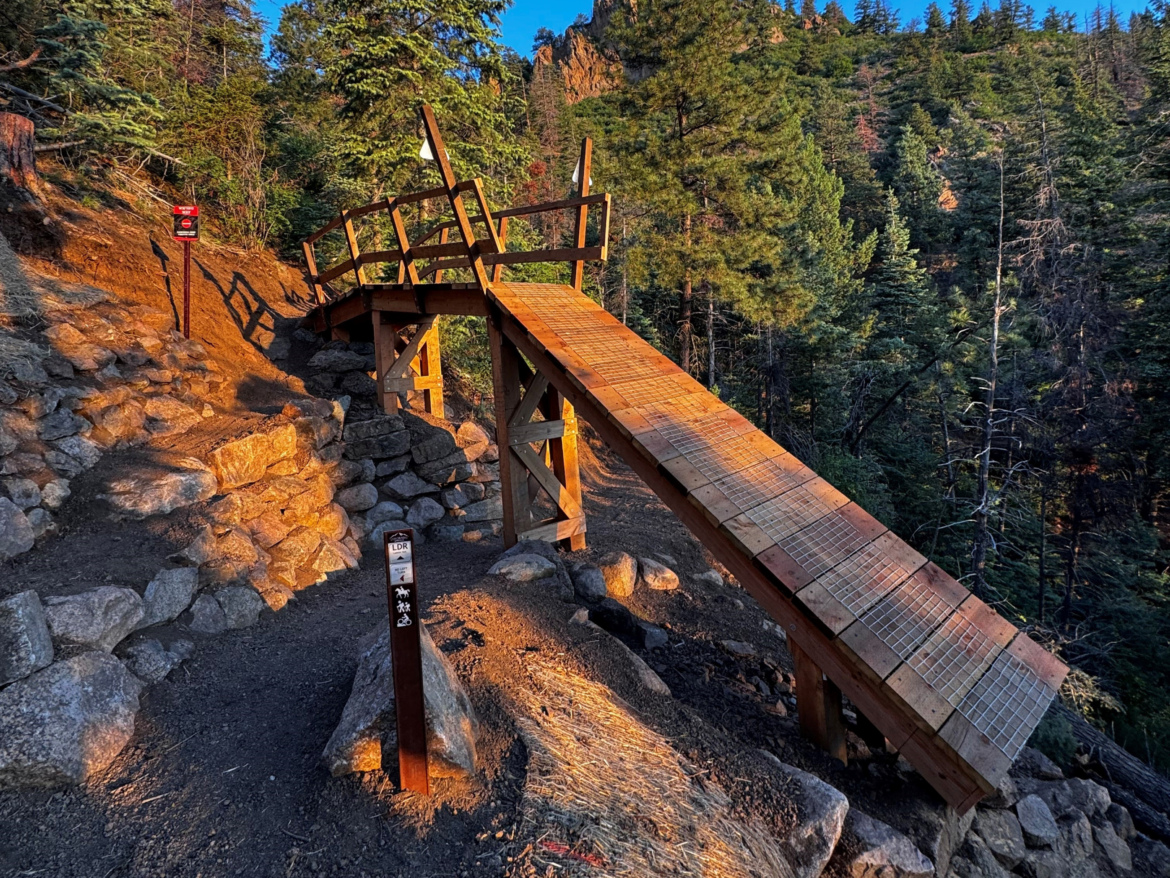 A wooden bridge crosses a rocky path in a forested area, surrounded by tall trees. The bridge is sunlit, highlighting its structure, while nearby trail signs indicate the direction or rules for hikers. The landscape features a mixture of rocky terrain and dirt paths, with shadows creating a sense of depth. Captain Morgan mountain bike trail.