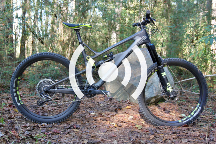 A mountain bike with a sleek gray frame and vibrant green accents, positioned on a bed of leaves in a forested area. The bike features thick tires designed for rugged terrain and is equipped with a front suspension fork. In the background, tall trees and underbrush create a natural setting.