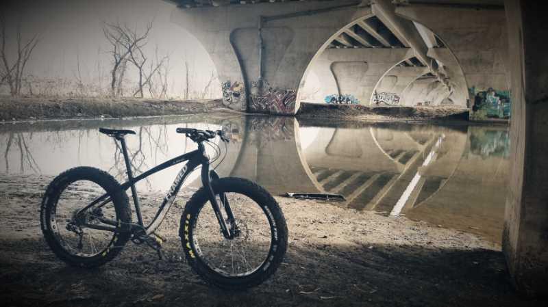 A black fat bike resting on the muddy bank of a river, with reflections visible in the water. The scene is set under a concrete overpass adorned with colorful graffiti, and barren trees are seen in the background. The atmosphere is moody and slightly foggy. Mendota/Ft Snelling mountain bike trail.