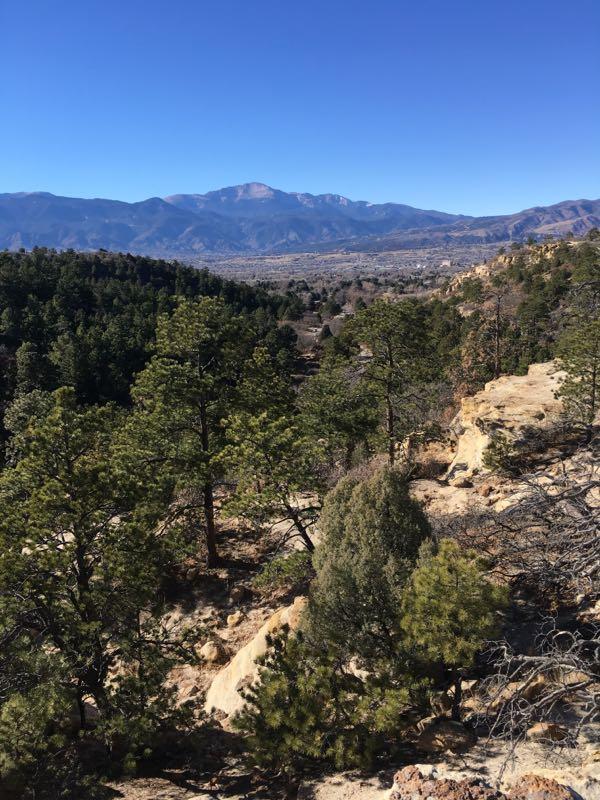 Scenic view of a mountainous landscape with lush green trees in the foreground, rocky terrain on the right, and distant mountains under a clear blue sky. Palmer Park mountain bike trail.