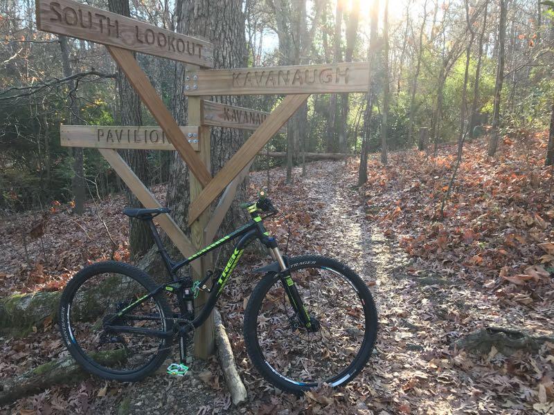 A mountain bike rests beside a wooden trail signpost in a forested area, indicating directions to "South Lookout," "Kavanaugh," and "Pavilion." Sunlight filters through the trees, casting a warm glow on the leaf-strewn ground and the bike. Allsopp Park mountain bike trail.