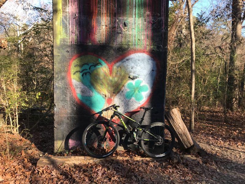 A black mountain bike leaning against a graffiti-covered wall featuring a colorful heart design, surrounded by trees and fallen leaves in a wooded area. Allsopp Park mountain bike trail.