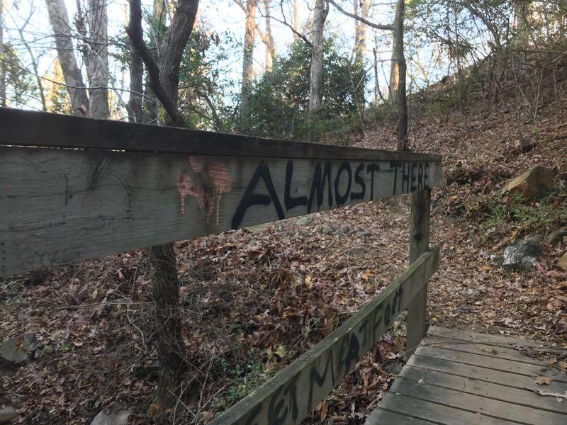 Wooden railing along a forest path, featuring graffiti that reads "ALMOST THERE." The scene is surrounded by trees and scattered autumn leaves, with a sloped, earthy path leading further into the woods. Allsopp Park mountain bike trail.