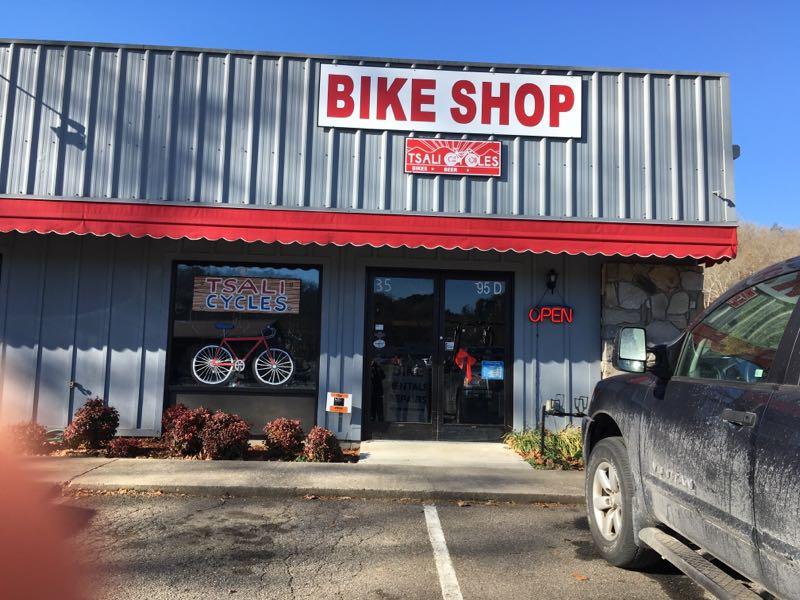 A bike shop with a gray exterior and a red awning, featuring a sign that reads "BIKE SHOP" prominently at the top. The storefront displays the name "Tsali Cycles" and has a window showcasing a bicycle. An "OPEN" sign is visible in the door, and a parked vehicle is seen nearby. Bright sunlight illuminates the scene, with some plants in front of the shop.