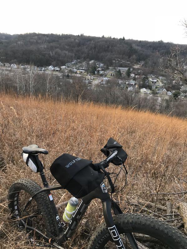 A mountain bike parked in a field of tall, dry grass, overlooking a distant residential area with trees on the hillside in the background. Memorial Park mountain bike trail.