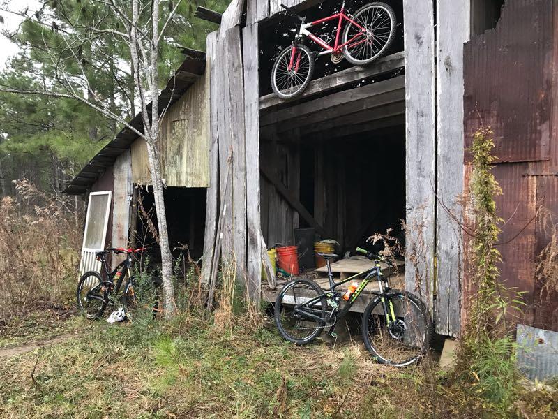 An old wooden barn surrounded by overgrown grass and trees, featuring two bicycles parked outside and a third bicycle hanging above in the loft. Inside the barn, there are various items, including buckets and tools, visible in the dim light. Mt. Zion Bike Trails mountain bike trail.
