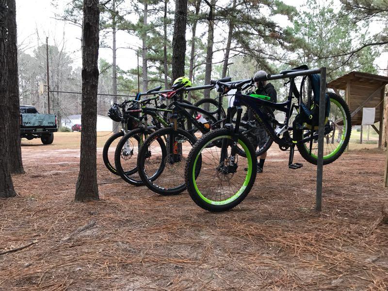 A row of mountain bikes hanging on a bike rack in a wooded outdoor area, with two cyclists nearby. The setting features tall trees and a dirt ground covered in pine needles. A parked truck is visible in the background. Mt. Zion Bike Trails mountain bike trail.