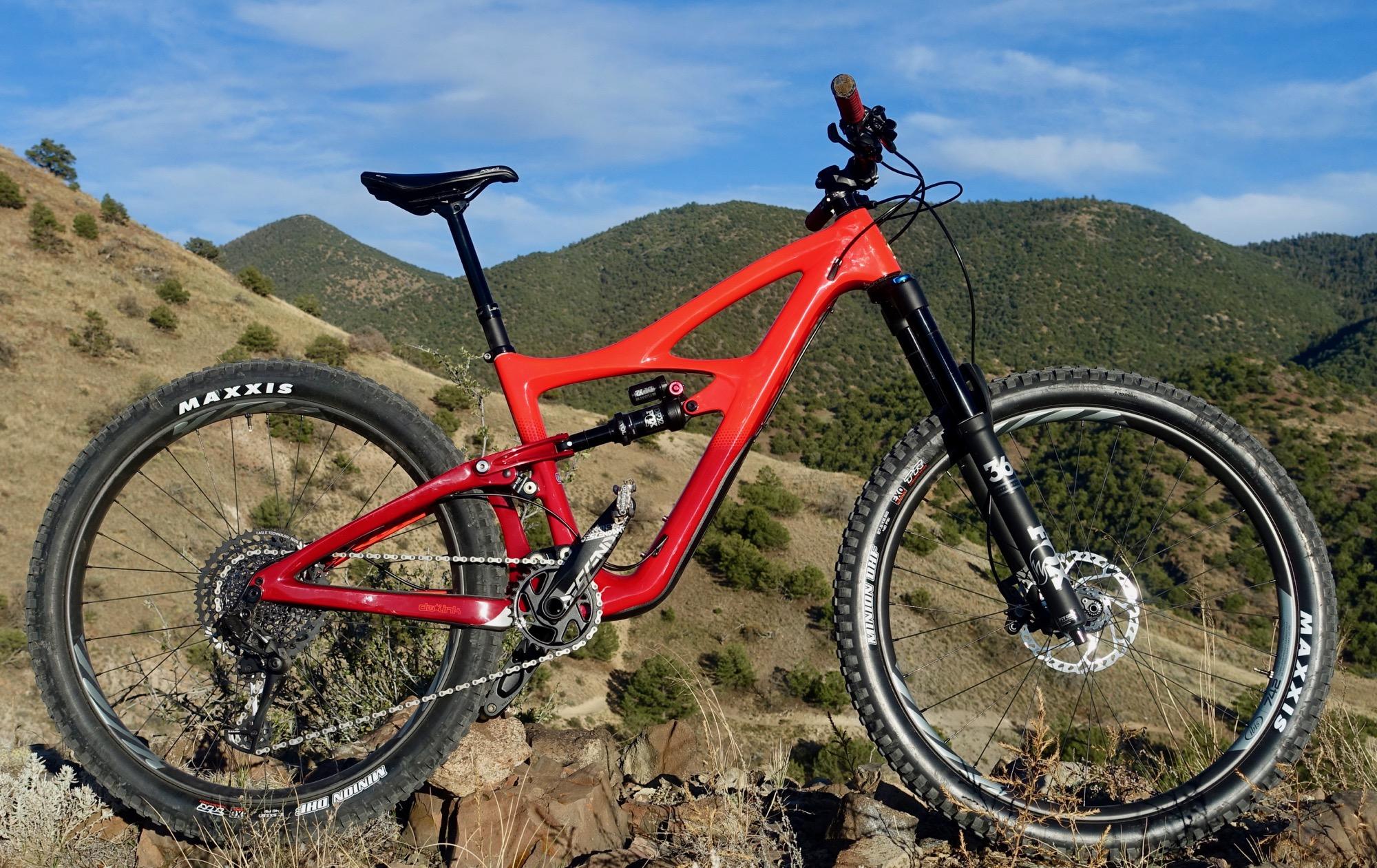 Ibis Mojo HD4: A red mountain bike parked on rocky terrain with scenic green hills and a blue sky in the background. The bike features thick tires marked with "MAXXIS" and a prominent suspension fork, indicating it's designed for off-road cycling.