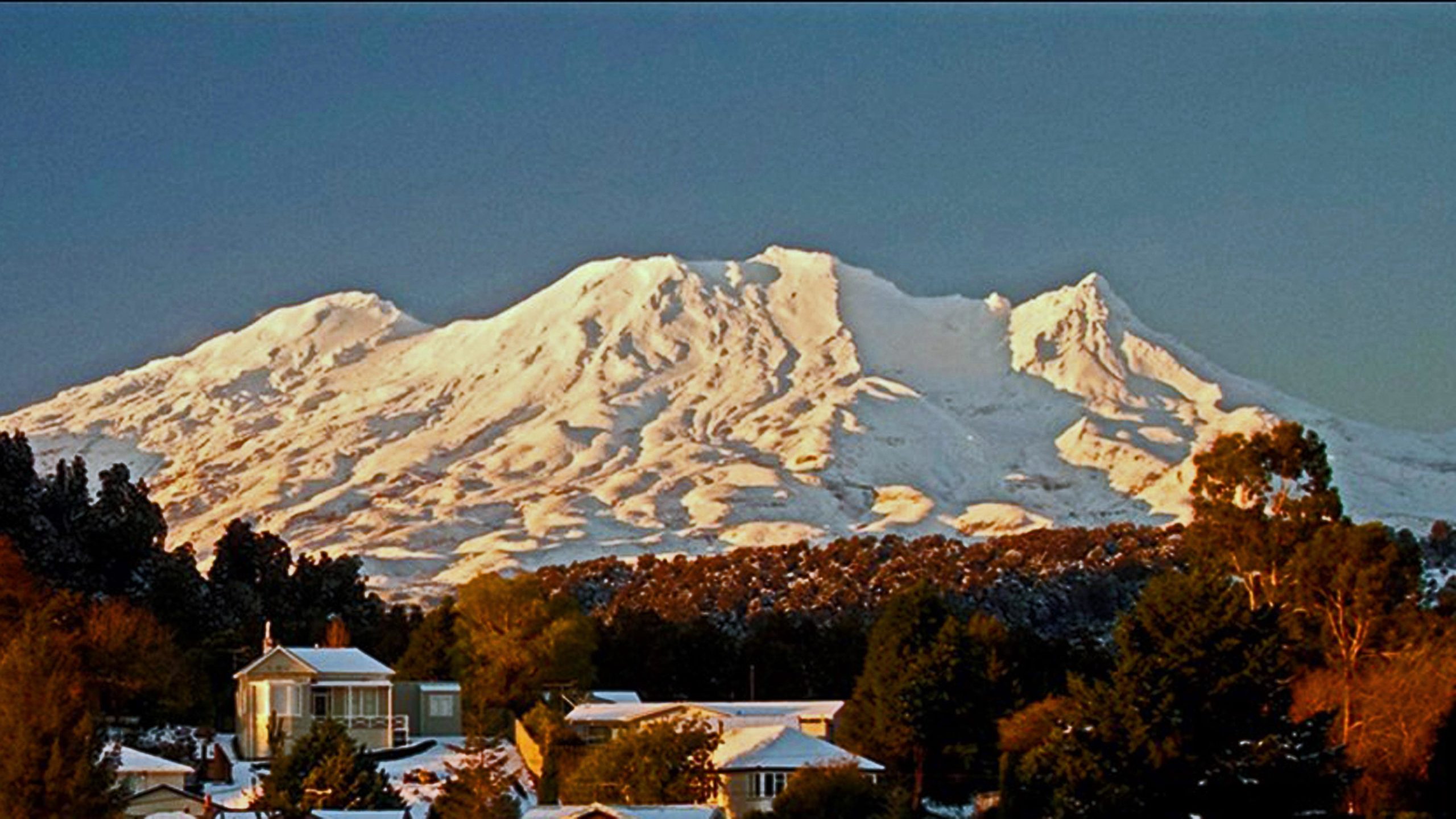 A scenic view of a snow-capped mountain range in the background, with a cluster of houses and trees in the foreground, set against a clear blue sky. The mountain displays gentle slopes and peaks, illuminated by soft sunlight, while the landscape below is adorned with patches of snow. Rangataua Forest mountain bike trail.