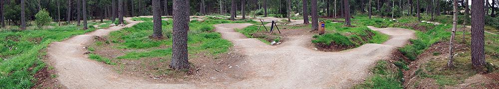 A winding dirt bike trail surrounded by trees, featuring multiple curves and turns on a grassy landscape. Tarland Trails mountain bike trail.