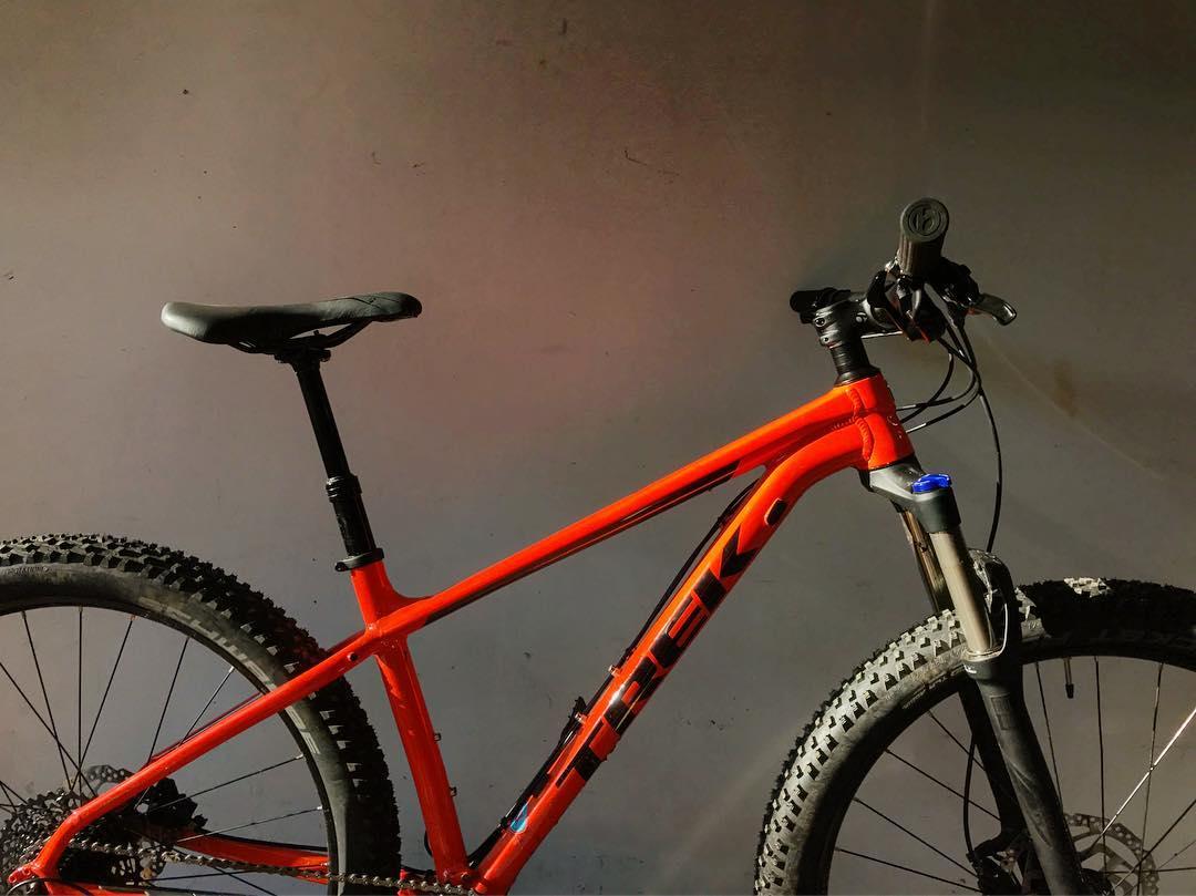 Trek Roscoe 8: A close-up view of a bright orange mountain bike with a sleek design, featuring a prominent Trek logo on the frame. The bike has thick, textured tires suitable for rough terrain, and a basic fork, with visible gear components. The background is a neutral gray, highlighting the bike's vibrant color.