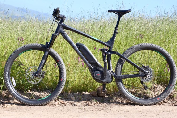 A black electric mountain bike parked on a dirt pathway surrounded by tall green grass and wildflowers. The bike features wide tires, a visible battery, and a sleek design, suitable for off-road biking.