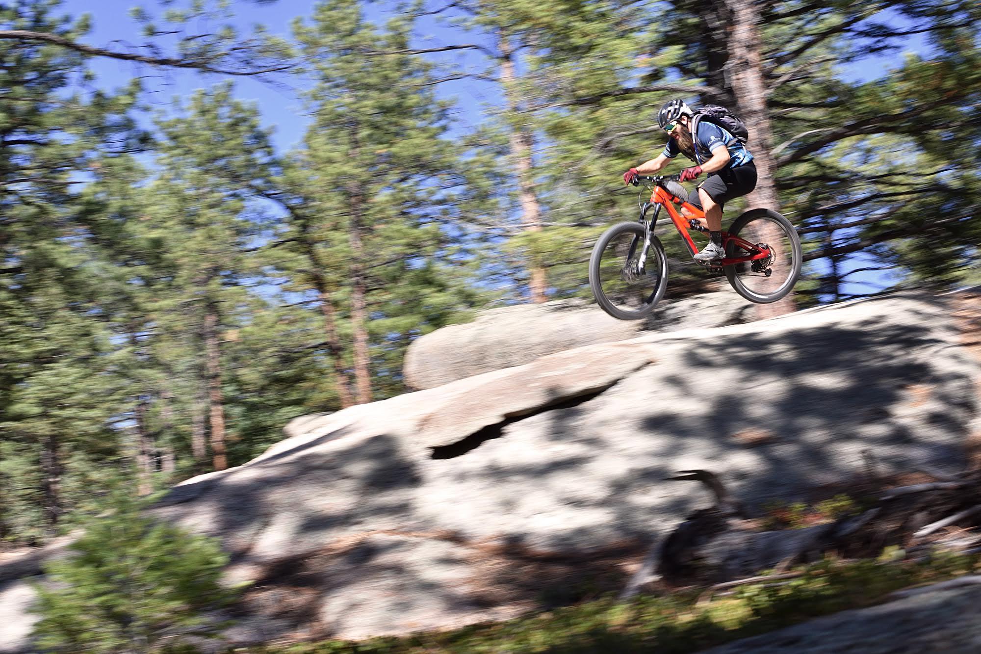 Ibis Mojo HD4: A mountain biker in motion jumping off a rocky outcrop, surrounded by tall trees in a forested area. The image captures a sense of speed and adventure.