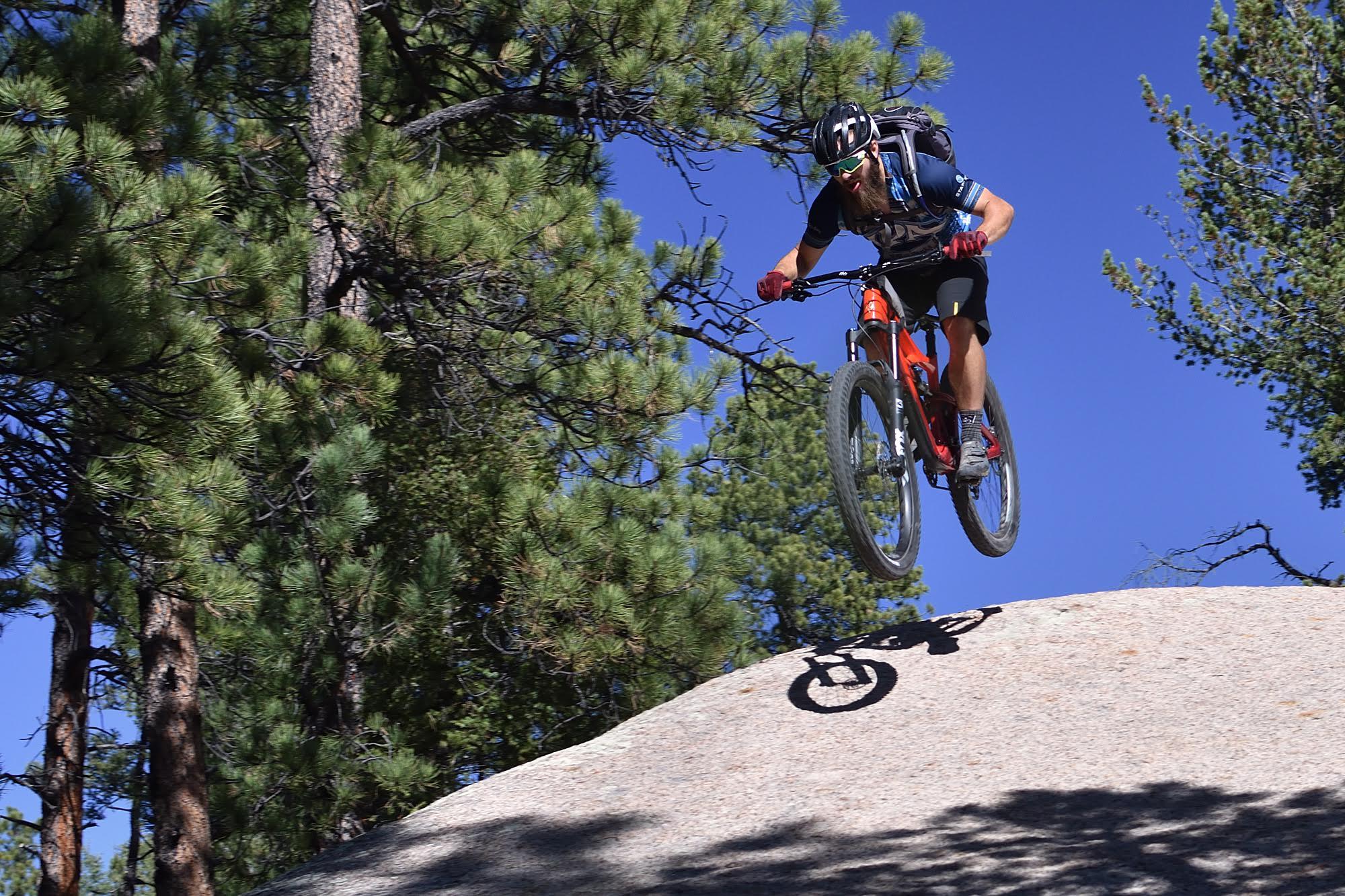 Ibis Mojo HD4: A mountain biker performing a jump off a rock in a forested area, surrounded by pine trees and a clear blue sky. The biker wears a helmet and protective gear, showcasing an action-packed moment in outdoor sports.
