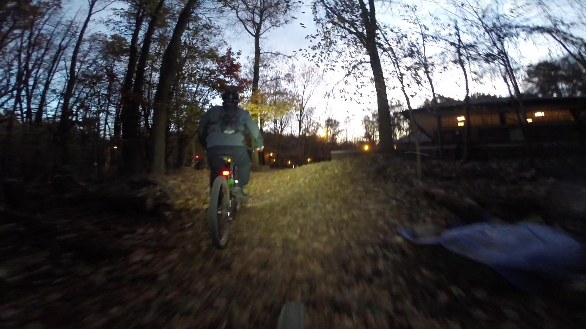 A person riding a mountain bike on a leaf-covered trail in a wooded area during dusk. The scene features trees shedding autumn leaves and a faint glow from lights in a nearby structure. Richmond Avenue and Forest Hill road mountain bike trail.