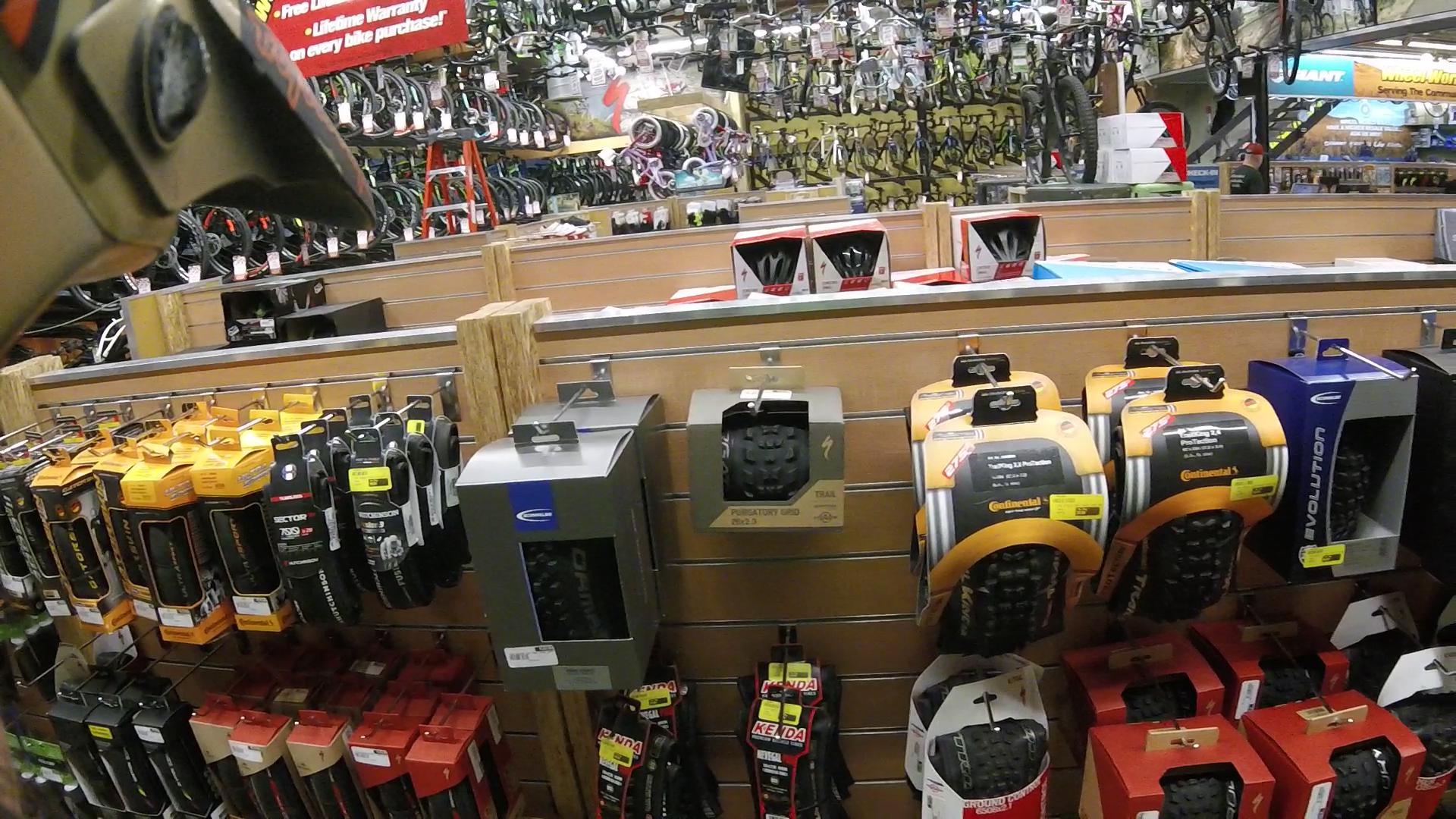 A display of bicycle tires and accessories in a bike shop, featuring various brands and types of tires, hanging on shelves. Background shelves are stocked with additional bicycles and equipment, creating a busy retail environment.
