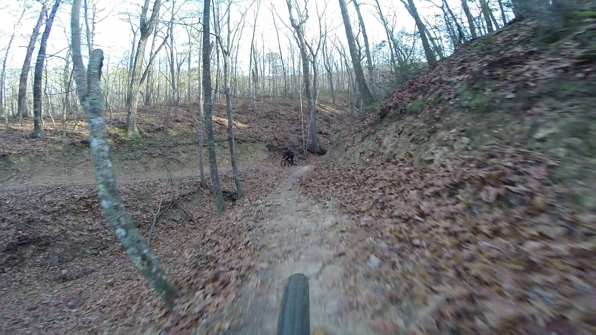 A mountain biker rides along a winding dirt trail surrounded by trees in a forest setting, with autumn leaves covering the ground. The image captures the perspective from the bike, showcasing a sloped path and the natural, wooded environment. Carvin's Cove Trail system mountain bike trail.