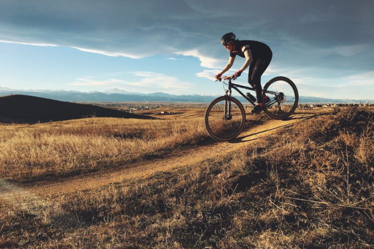 A cyclist navigating a dirt trail on a mountain bike, set against a backdrop of rolling hills and a cloudy sky. Snow-capped mountains are visible in the distance, and the landscape is predominantly dry with golden grass. The cyclist is wearing a helmet and athletic clothing, emphasizing an active outdoor lifestyle.