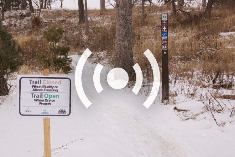 Signage indicating the status of a trail in a snowy landscape. One sign states "Trail Closed When Muddy or Above Freezing" and the other states "Trail Open When Dry or Frozen." A trail marker with icons for biking and hiking is also visible in the background, surrounded by trees and grass.