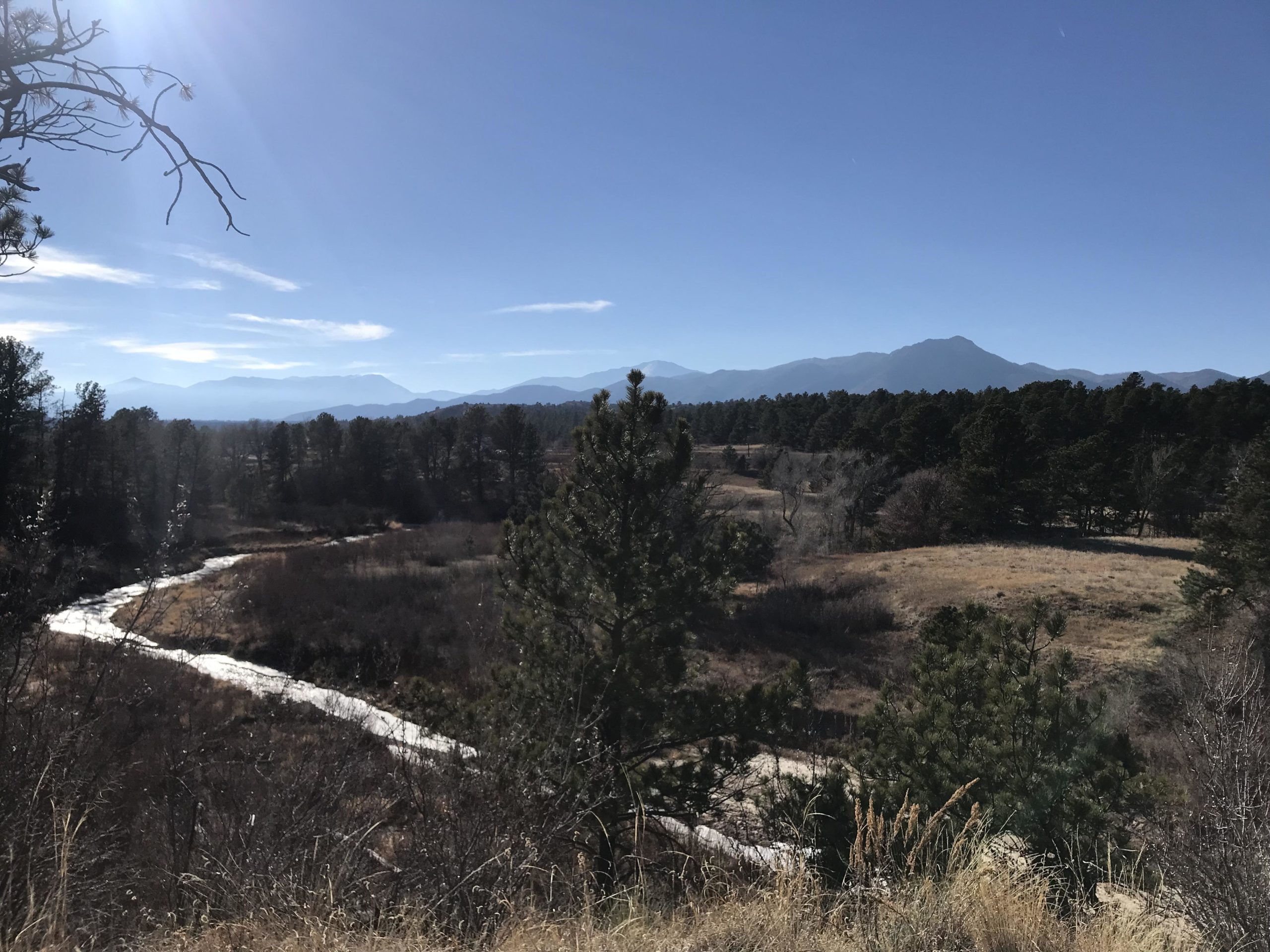 A scenic view of a landscape featuring a winding river, surrounded by trees and grassy fields, with mountains in the background under a clear blue sky. Santa Fe Trail mountain bike trail.