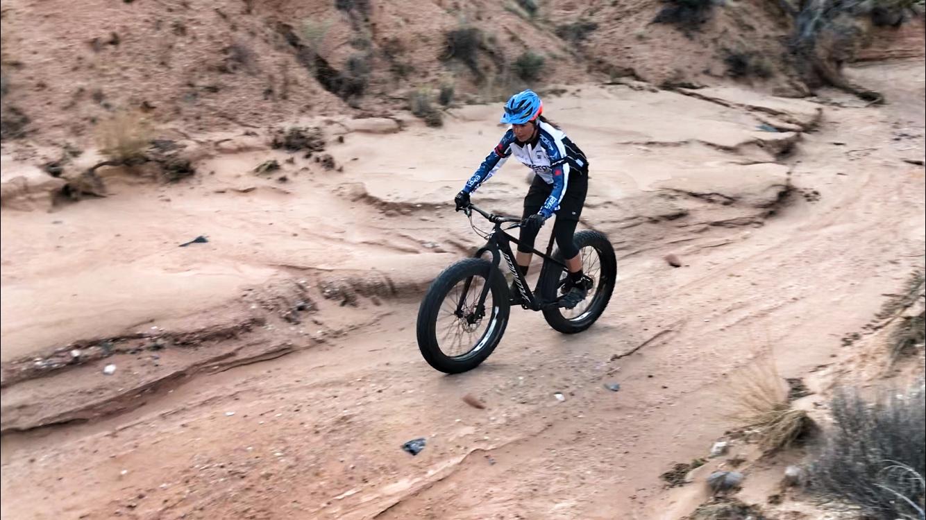 A cyclist riding a fat bike on a sandy, unpaved trail surrounded by rocky terrain and sparse vegetation. The cyclist is wearing a blue helmet and a long-sleeve jersey with colorful designs, focused on navigating the rough landscape. Mariposa Fat Bike Trails mountain bike trail.